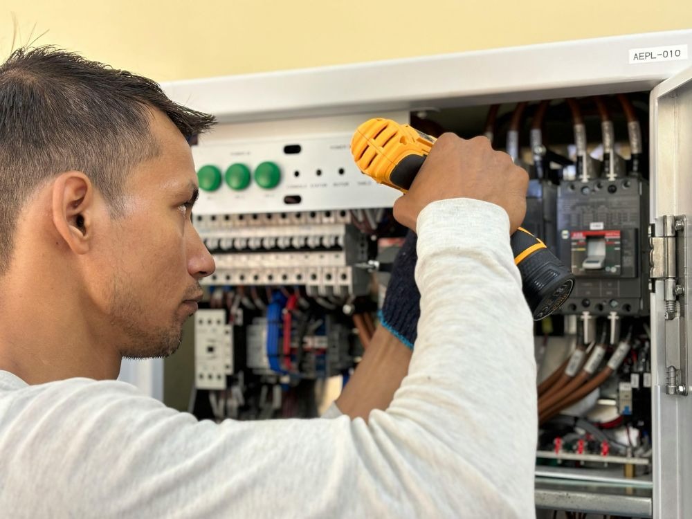 Um eletricista com luvas e uma chave de fenda elétrica amarela e preta trabalha em um painel de controle elétrico aberto. Ele está concentrado nos fios e disjuntores para consertar o problema.
