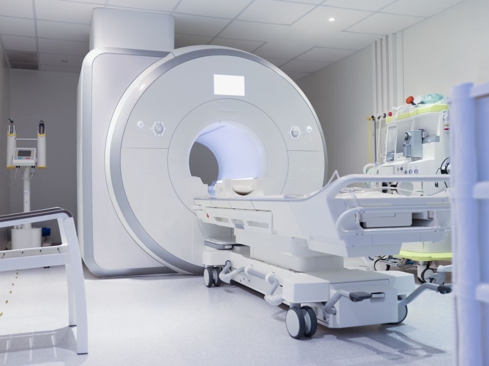 Uma máquina de ressonância magnética (MRI) grande e branca em uma sala de hospital limpa. A maca branca está posicionada para entrar no túnel do equipamento, que exige um sistema trifásico para operar.