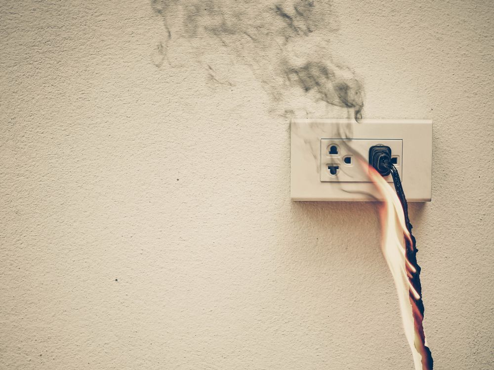 Curto circuito na tomada com cabo queimando e liberação de fumaça na parede, indicando falha grave na instalação elétrica.