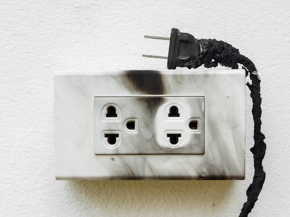 Tomada elétrica com marcas de queima e plugue derretido, ilustrando visualmente Como acontece o curto-circuito em instalações residenciais sobrecarregadas.