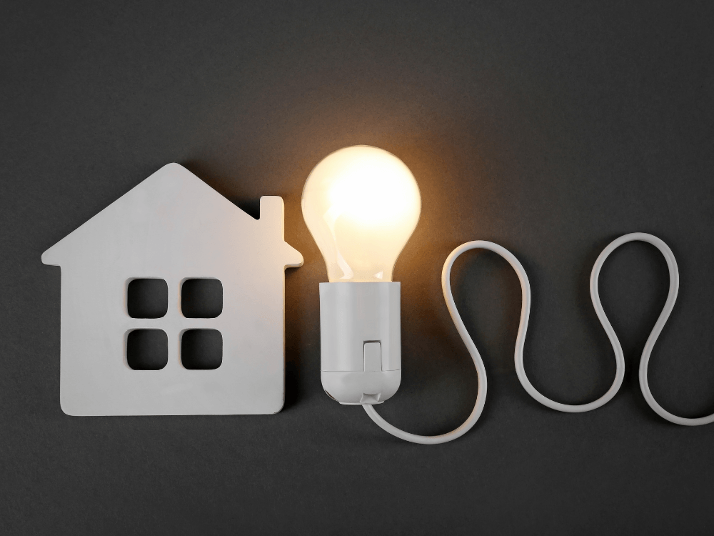 Lâmpada incandescente acesa ao lado de uma miniatura de casa branca de madeira com um cabo de energia sinuoso sobre fundo escuro demonstrando o que é a corrente elétrica.