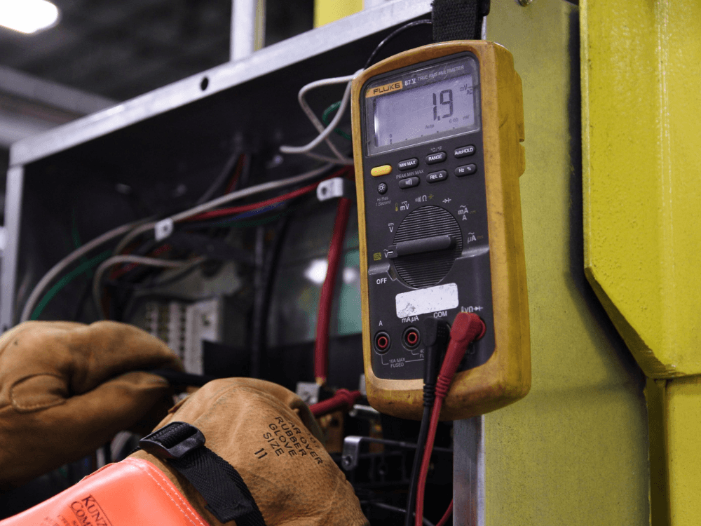 Multímetro digital conectado a componentes de um painel elétrico durante uma medição realizada por profissional com luvas de proteção.